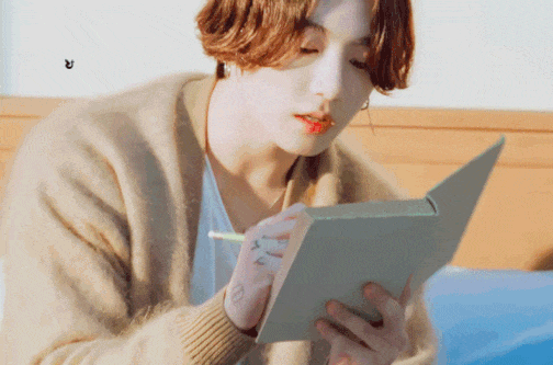 Gif of Jungkook in a loose white shirt under a light brown cardigan. He is holding and writing in a book while he mouths words.