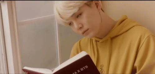 Gif of blond Suga in a saturated yellow hoodie, leaning against a glass paned window. He is holding an open red book.